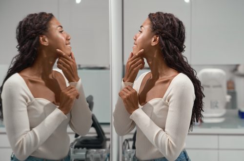 African woman smiles at a mirror while tracing the jawline with a fingertip, checking facial contours during a cosmetology consultation about skin firmness and definition.